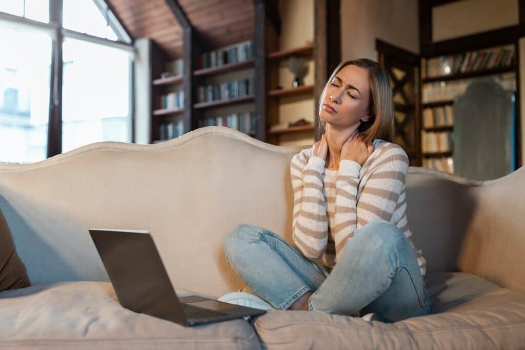 Femme fatiguée assise sur un canapé avec un ordinateur, se massant le cou.