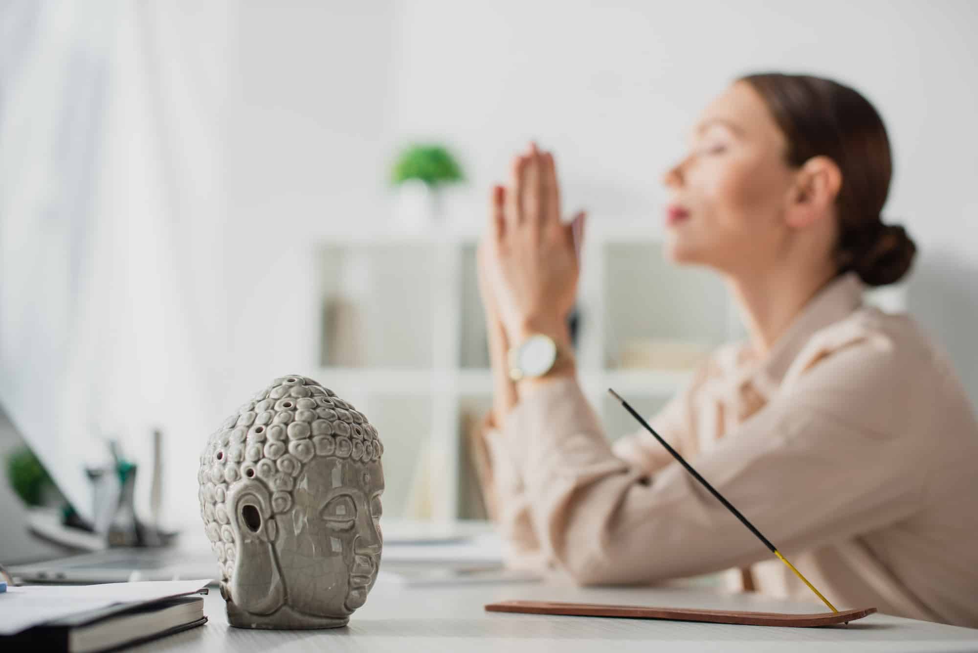 selective focus of businesswoman meditating with namaste gesture at workplace with Buddha head and